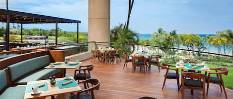 A restaurant deck with wooden tables and chairs, a large pillar, and a view of the ocean.