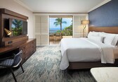 A hotel room with a bed, a TV, a desk, a chair, and a view of the ocean.