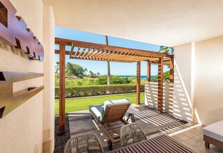 A covered outdoor seating area with a wooden pergola, lounge chairs, and a view of the ocean.