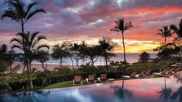 Una piscina infinita con sillones de descanso, palmeras y vista a la playa al atardecer.