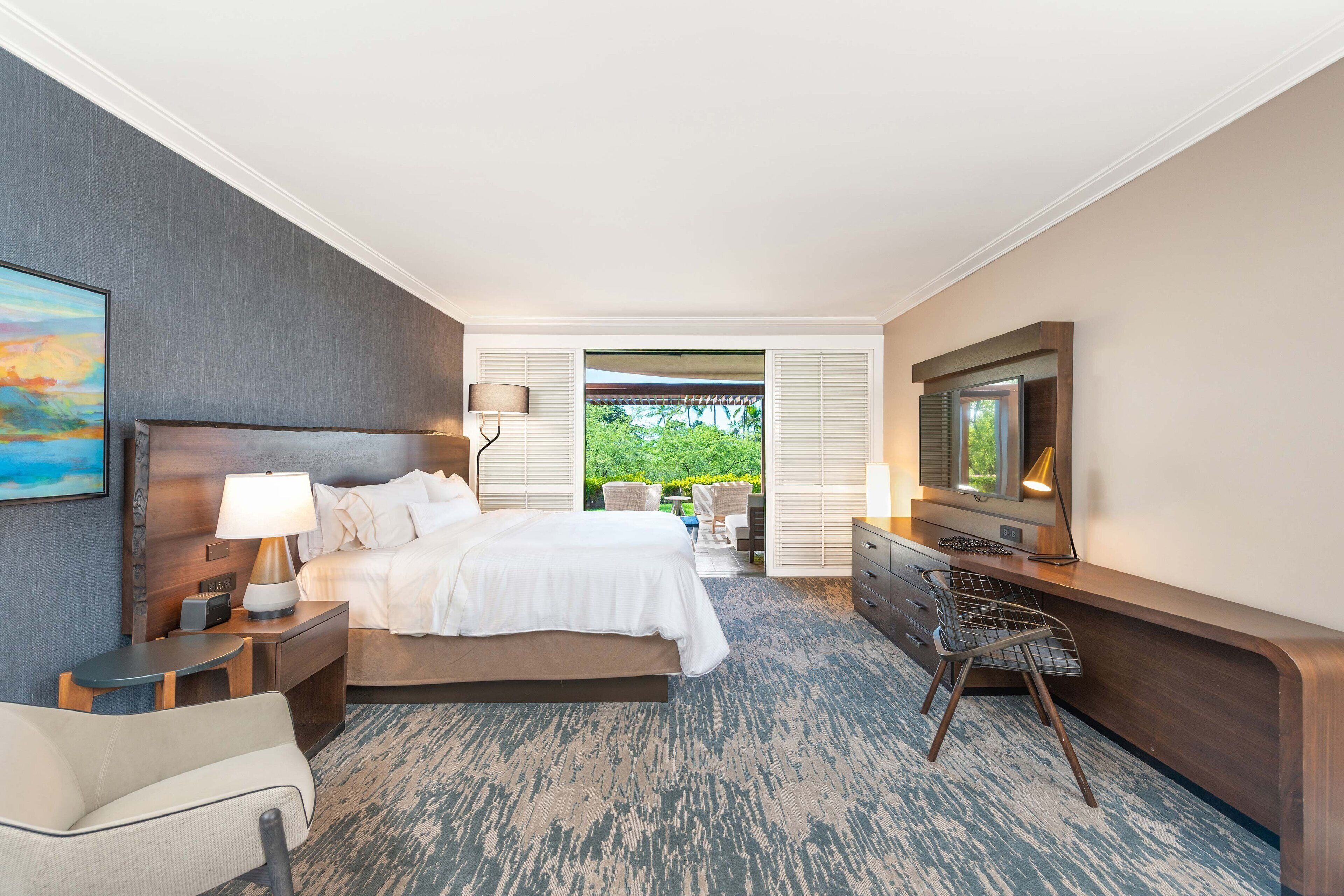 Habitación de hotel con una cama grande, un escritorio con una silla, un televisor y vistas a zonas verdes por la ventana.