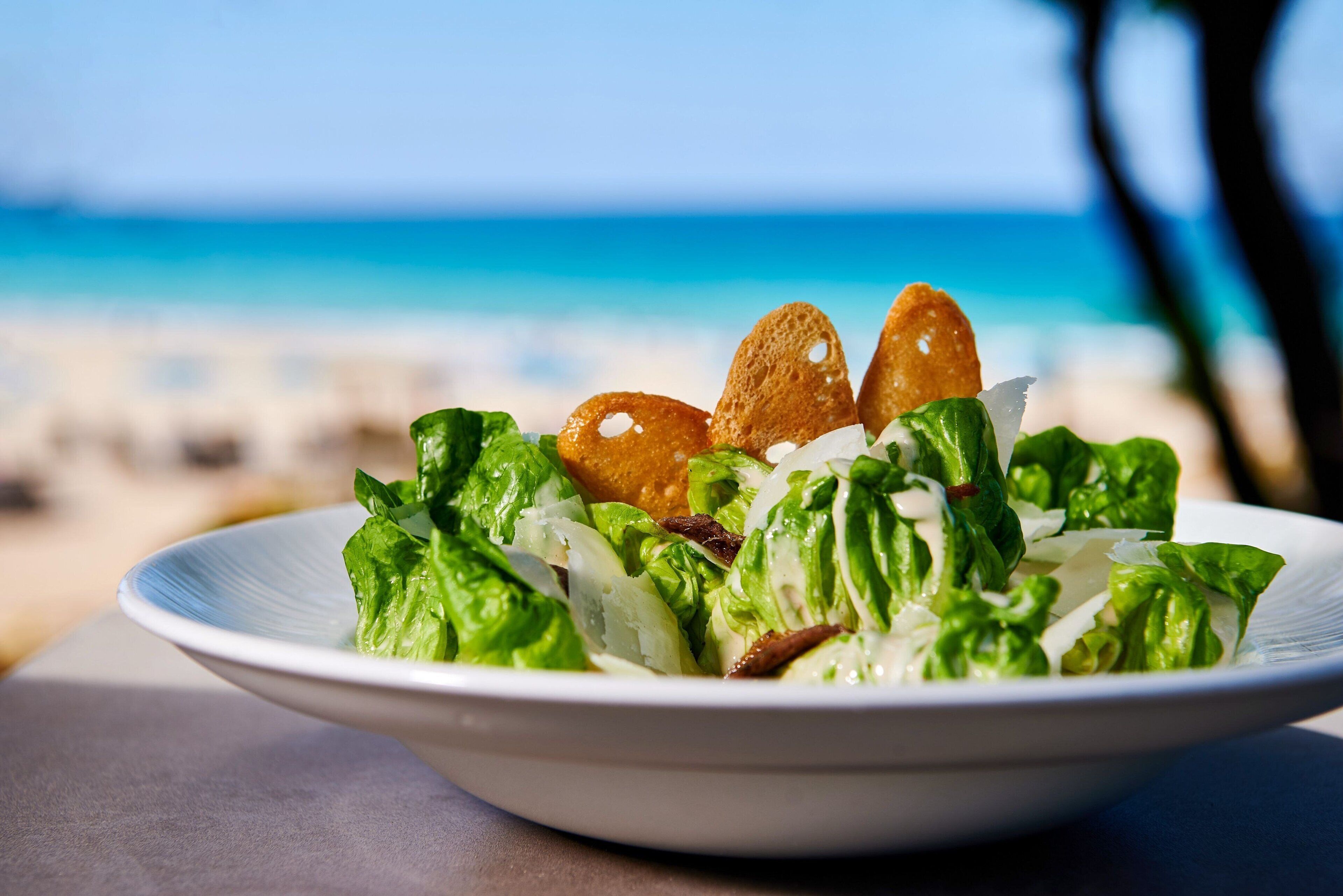 Un saladier rempli de salade avec des croûtons, posé sur une table, avec une vue sur la plage en arrière-plan.