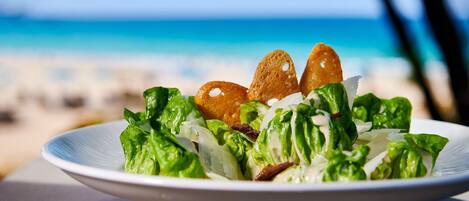 Un saladier rempli de salade avec des croûtons, posé sur une table, avec une vue sur la plage en arrière-plan.
