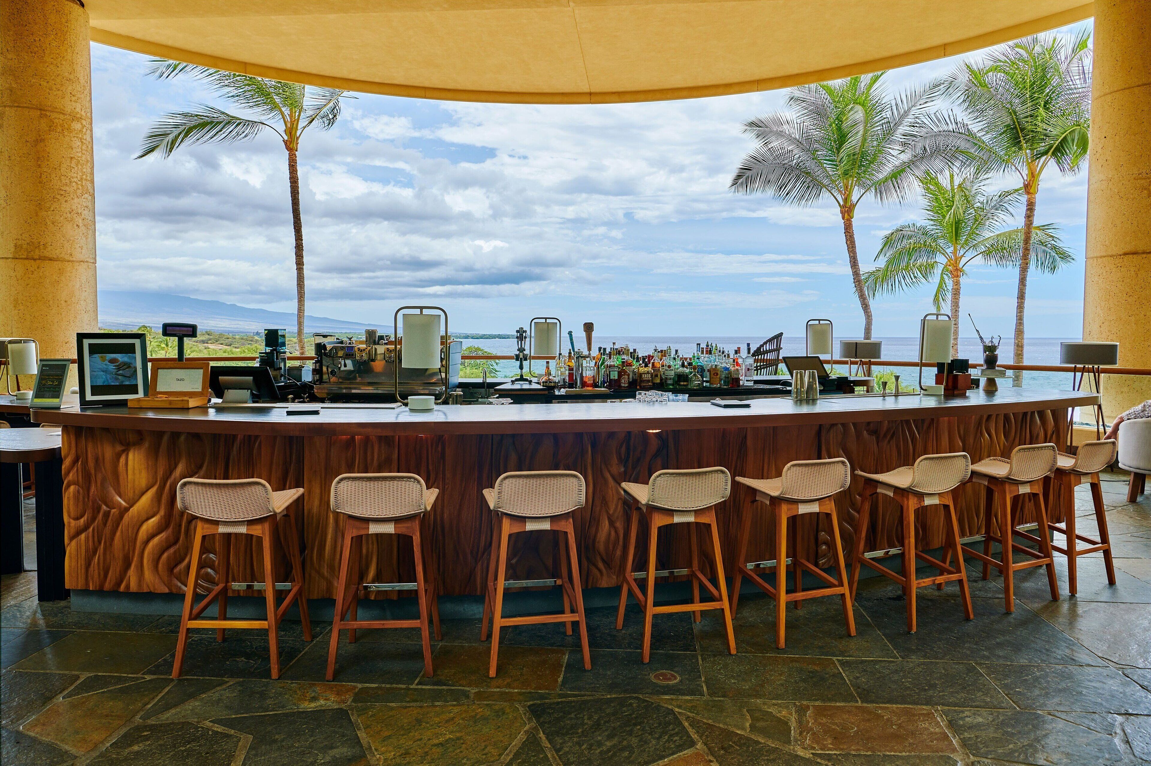 A bar with a view of palm trees and the ocean, wooden bar stools, and a stone-tiled floor.