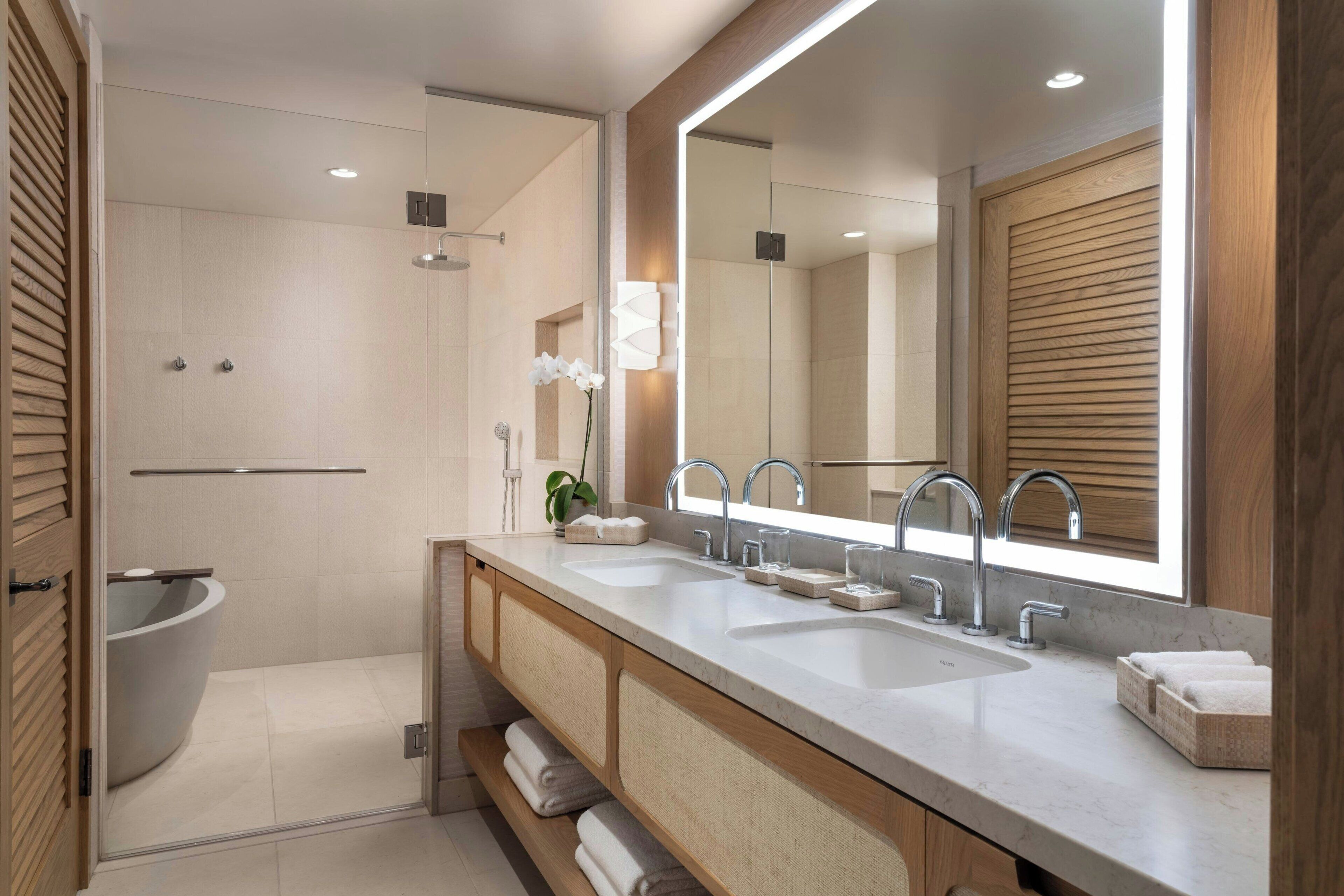 A modern bathroom with a large mirror, two sinks, a shower, and wooden cabinetry.