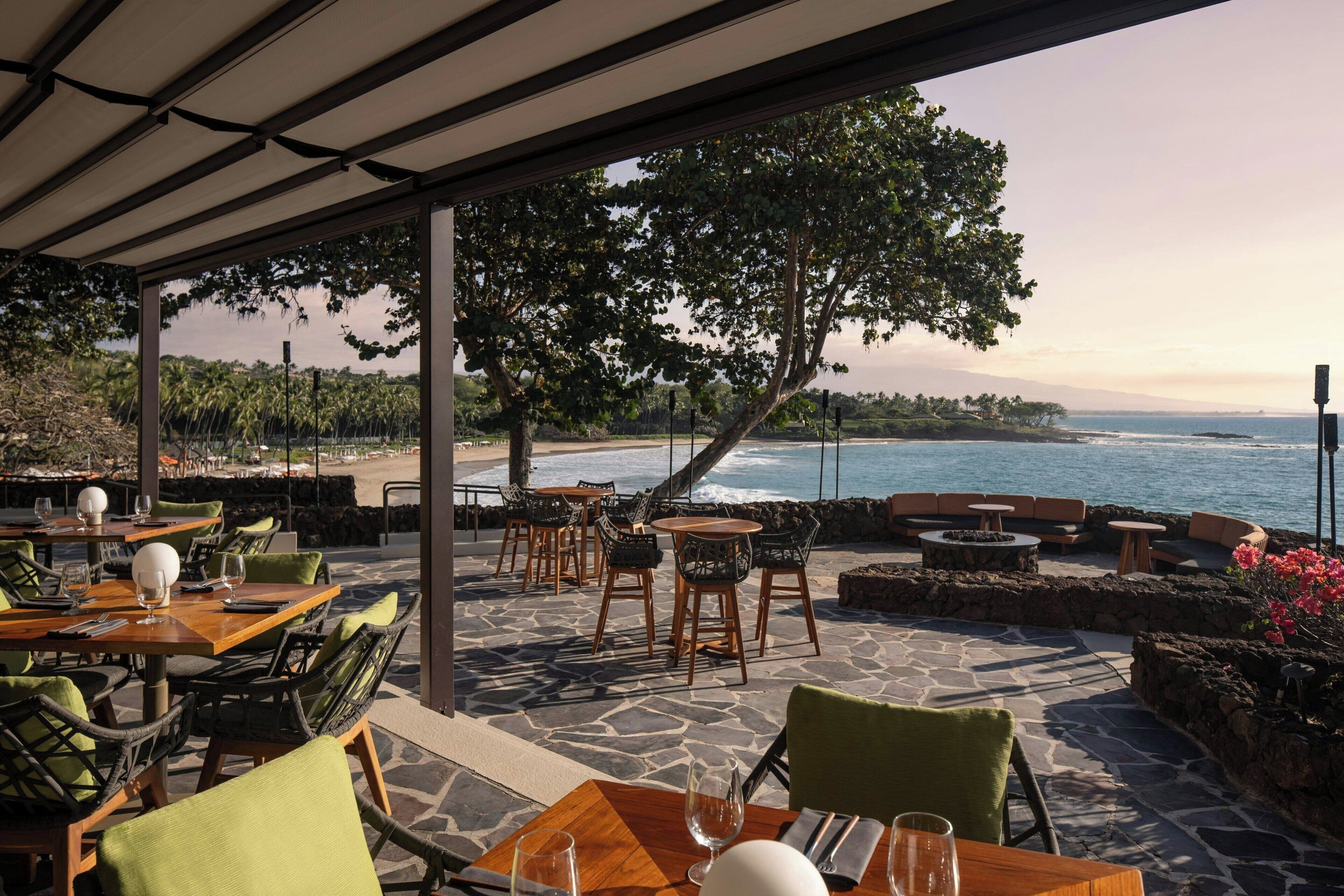 An outdoor dining area with tables and chairs, overlooking a beach and ocean.
