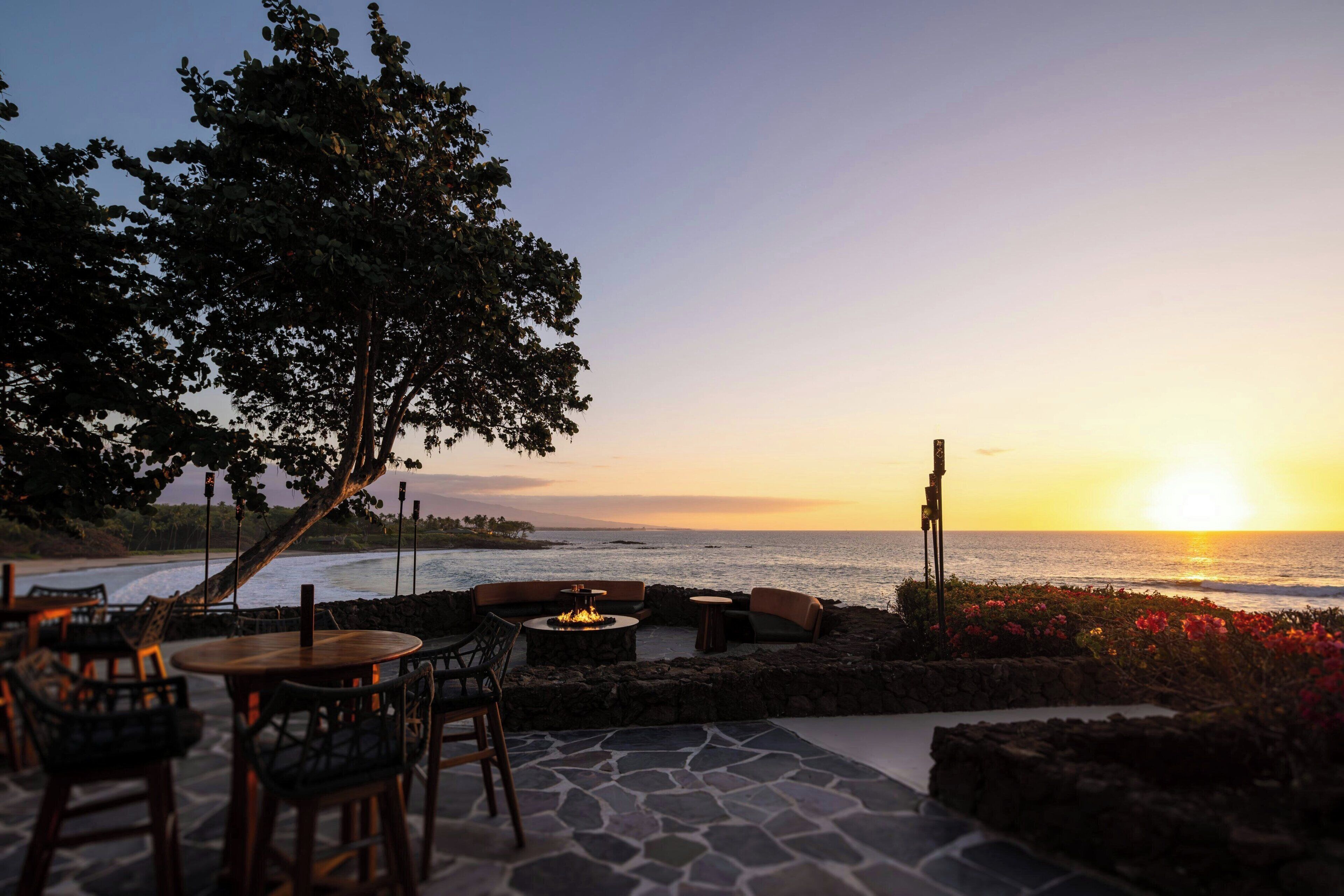 An outdoor seating area with stone flooring, wooden furniture, and a fire pit, overlooking the ocean at sunset.
