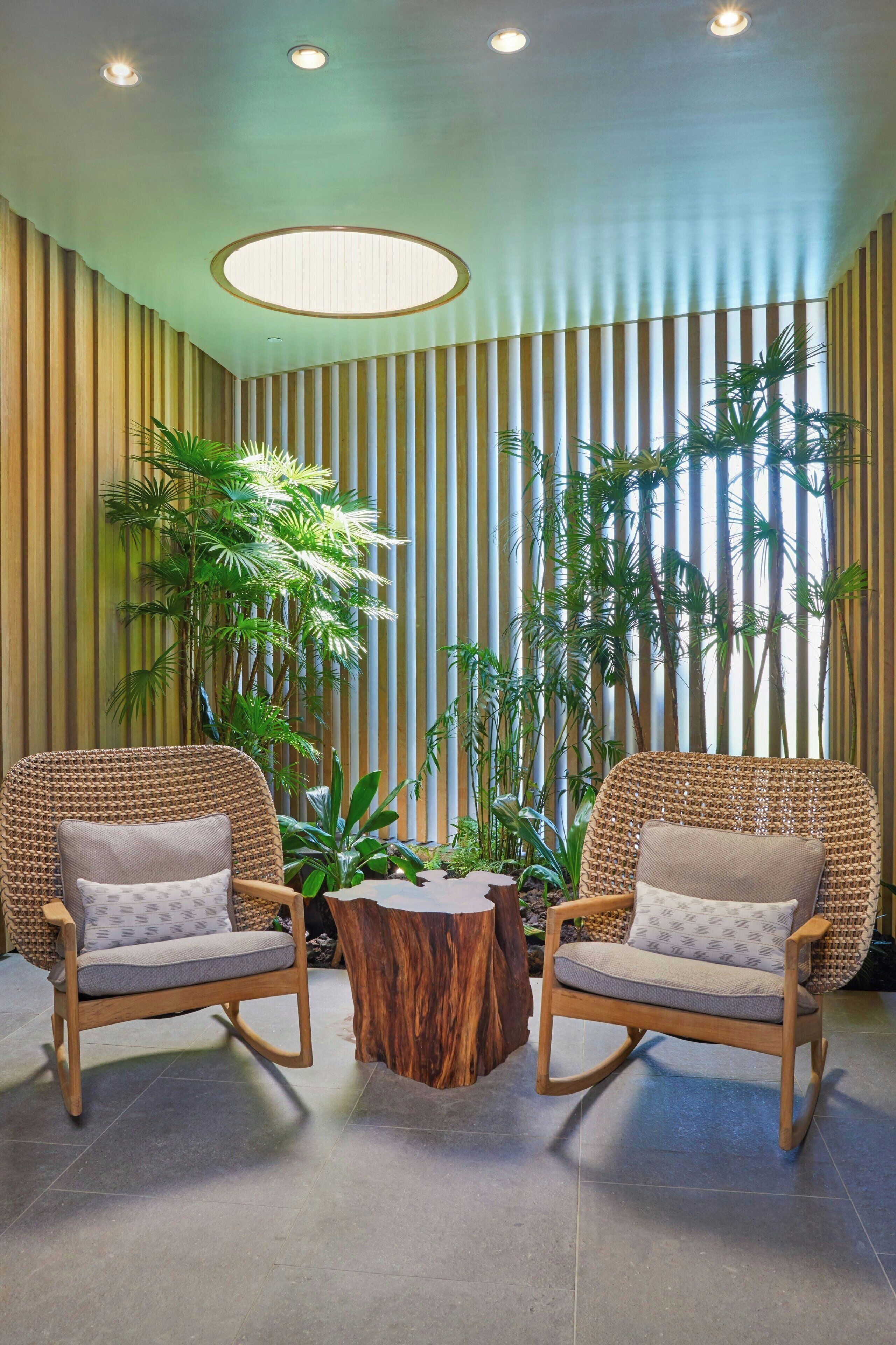 A room with two wicker chairs, a wooden coffee table, and tall plants against a striped wall.