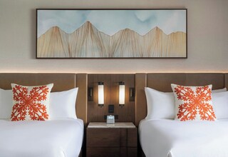 A hotel room with two beds, each with a white bedspread and a red floral pillow. There is a wooden headboard, a nightstand with a clock, and a wall-mounted artwork depicting mountains.