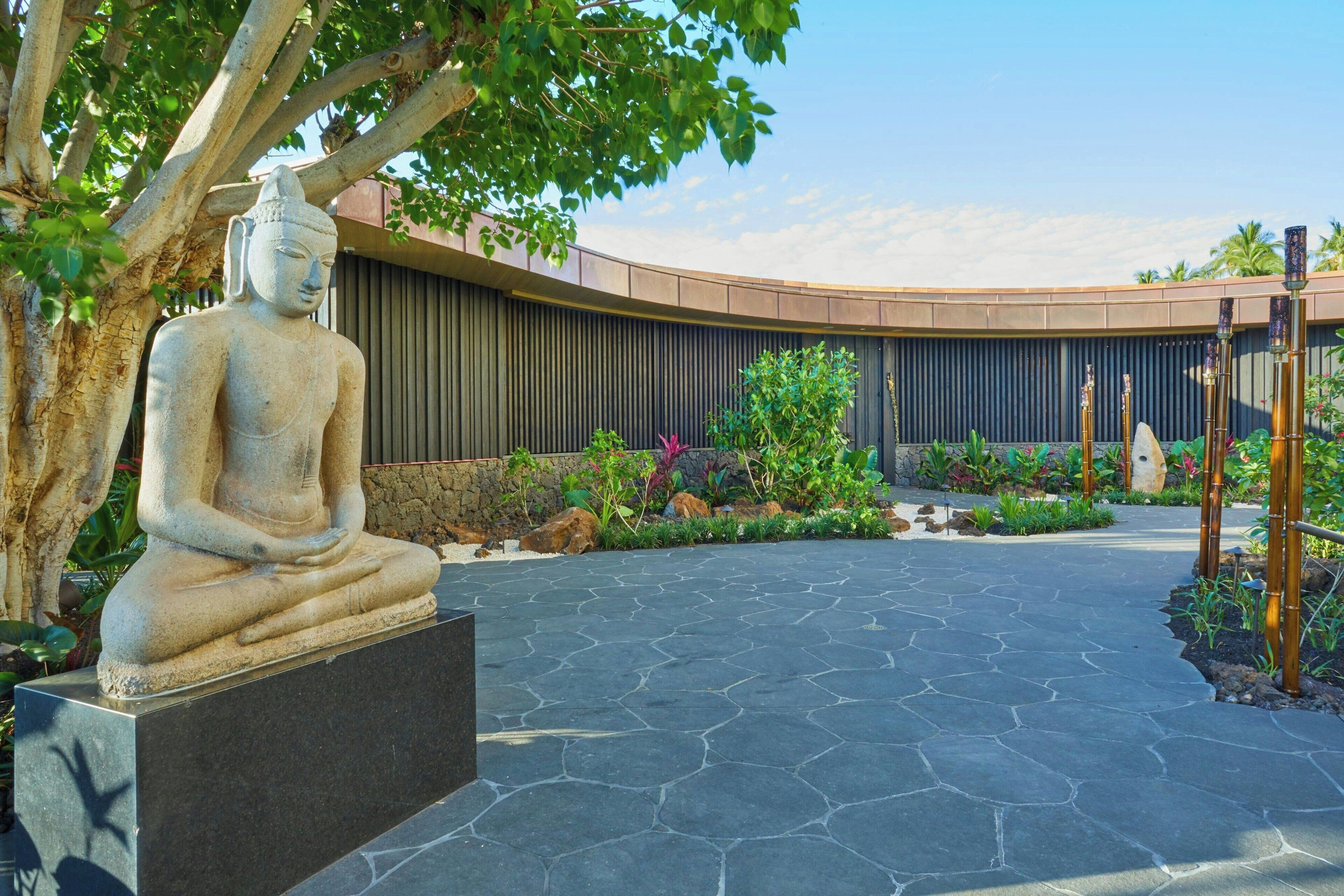 A stone Buddha statue on a pedestal, surrounded by a landscaped garden with a curved wall and a stone pathway.