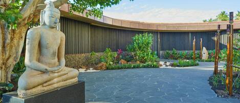 A stone Buddha statue on a pedestal, surrounded by a landscaped garden with a curved wall and a stone pathway.