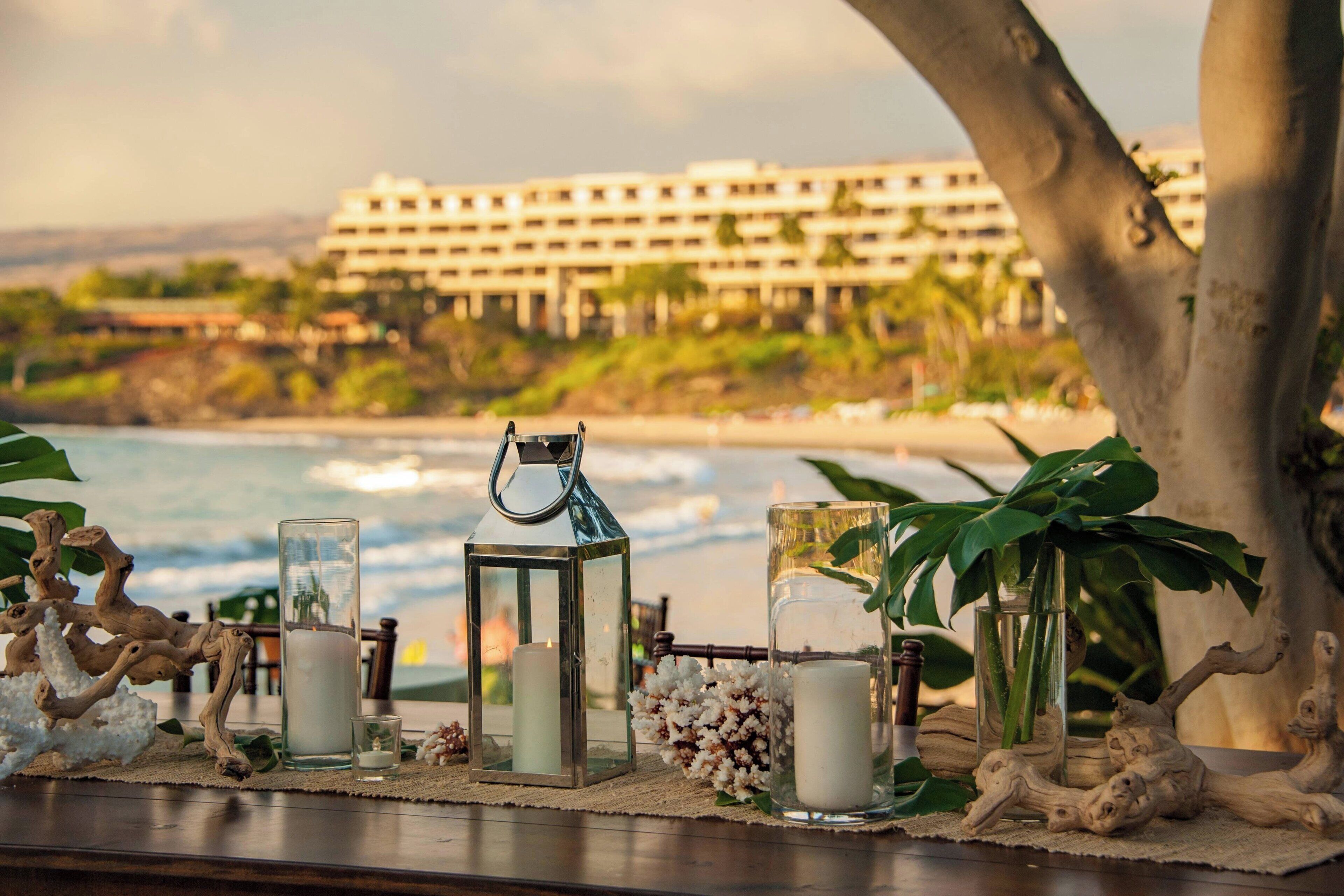 A table with candles, a lantern, and decorative items by a window overlooking a beach and hotel.