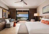 A hotel room with a bed, a sofa, a small table, a desk, a ceiling fan, a balcony with a view, and a painting on the wall.