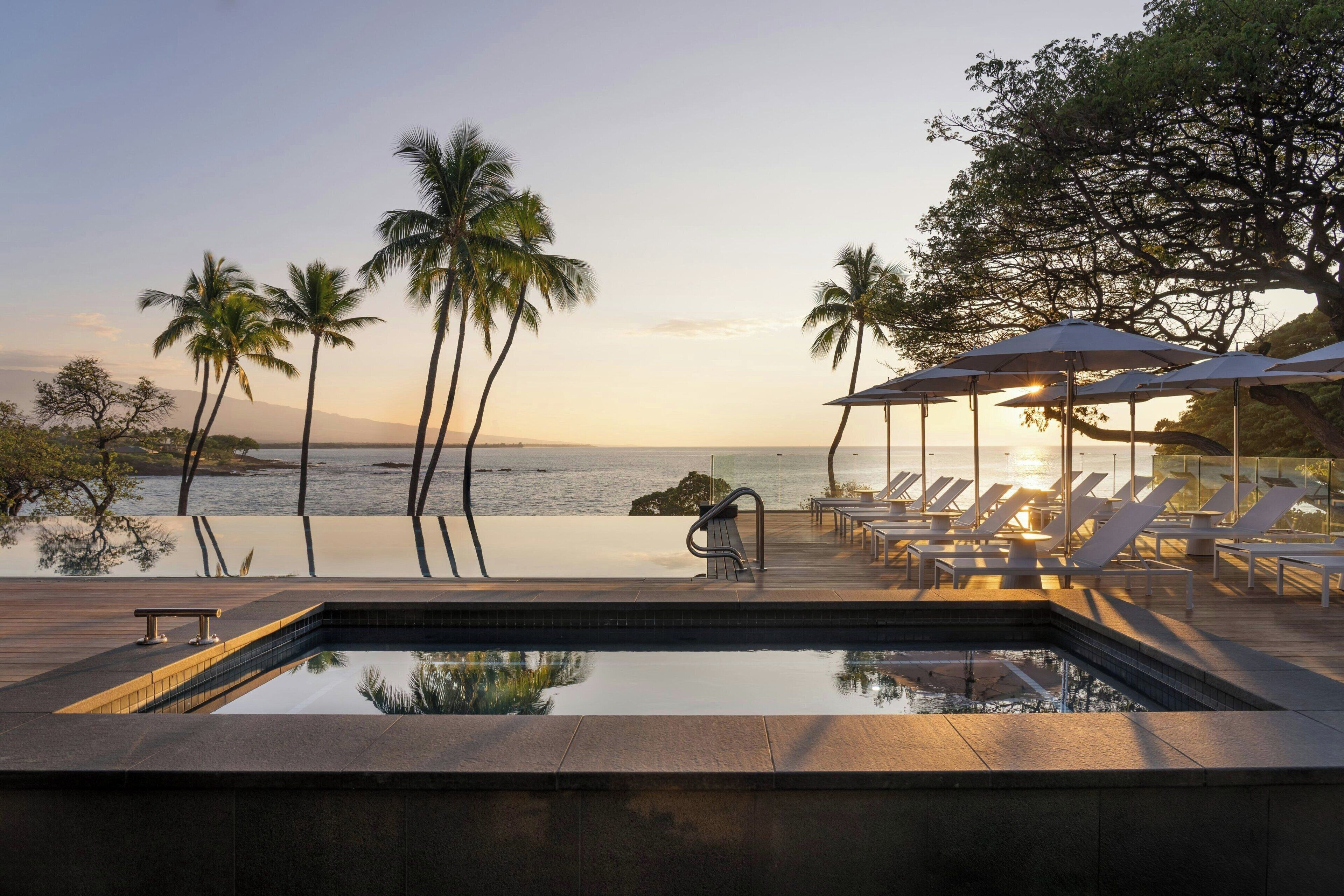 A poolside area with lounge chairs and umbrellas, overlooking a body of water and palm trees.