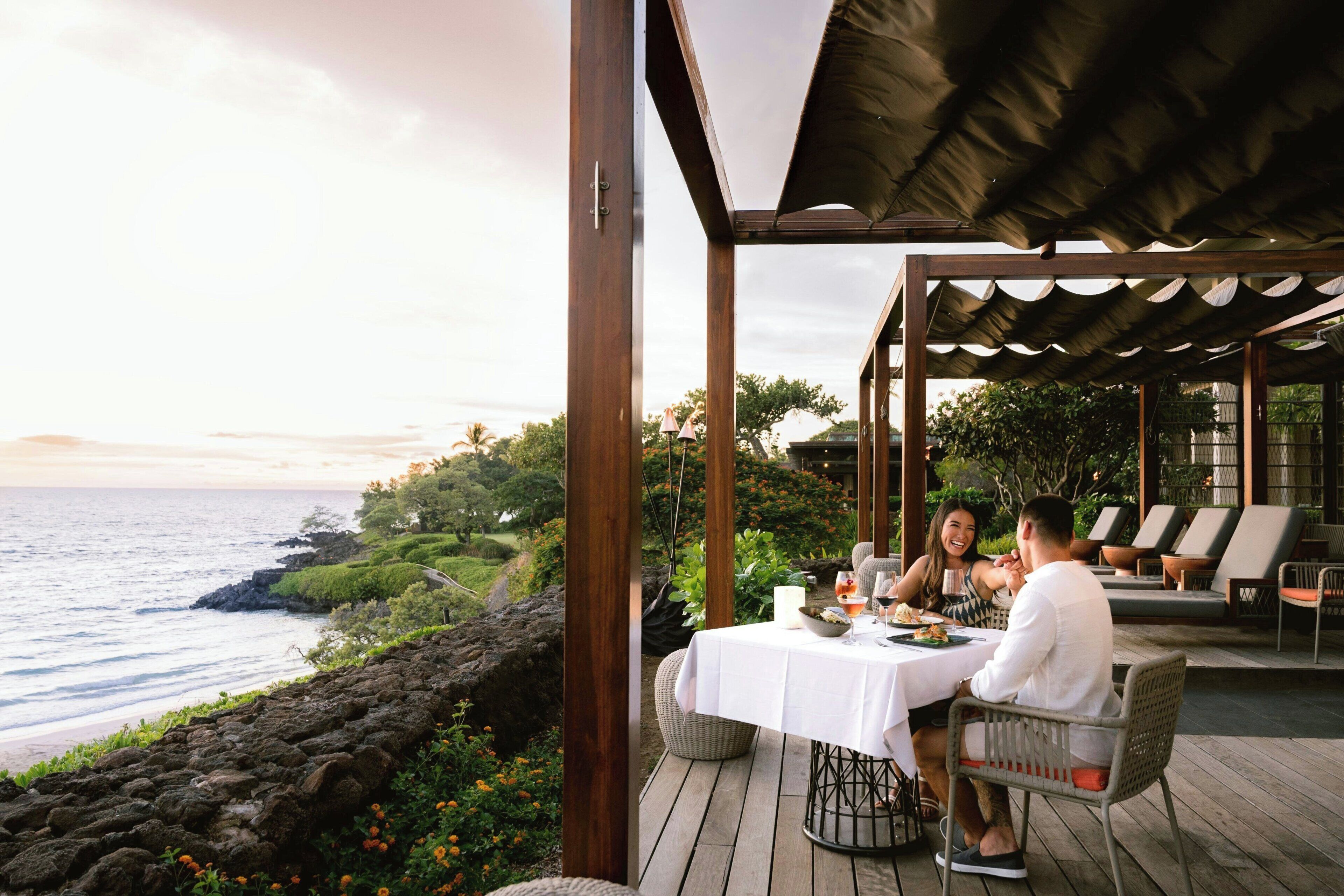 Une terrasse en bois avec une table dressée pour deux personnes, offrant une vue sur une côte rocheuse et l’océan.
