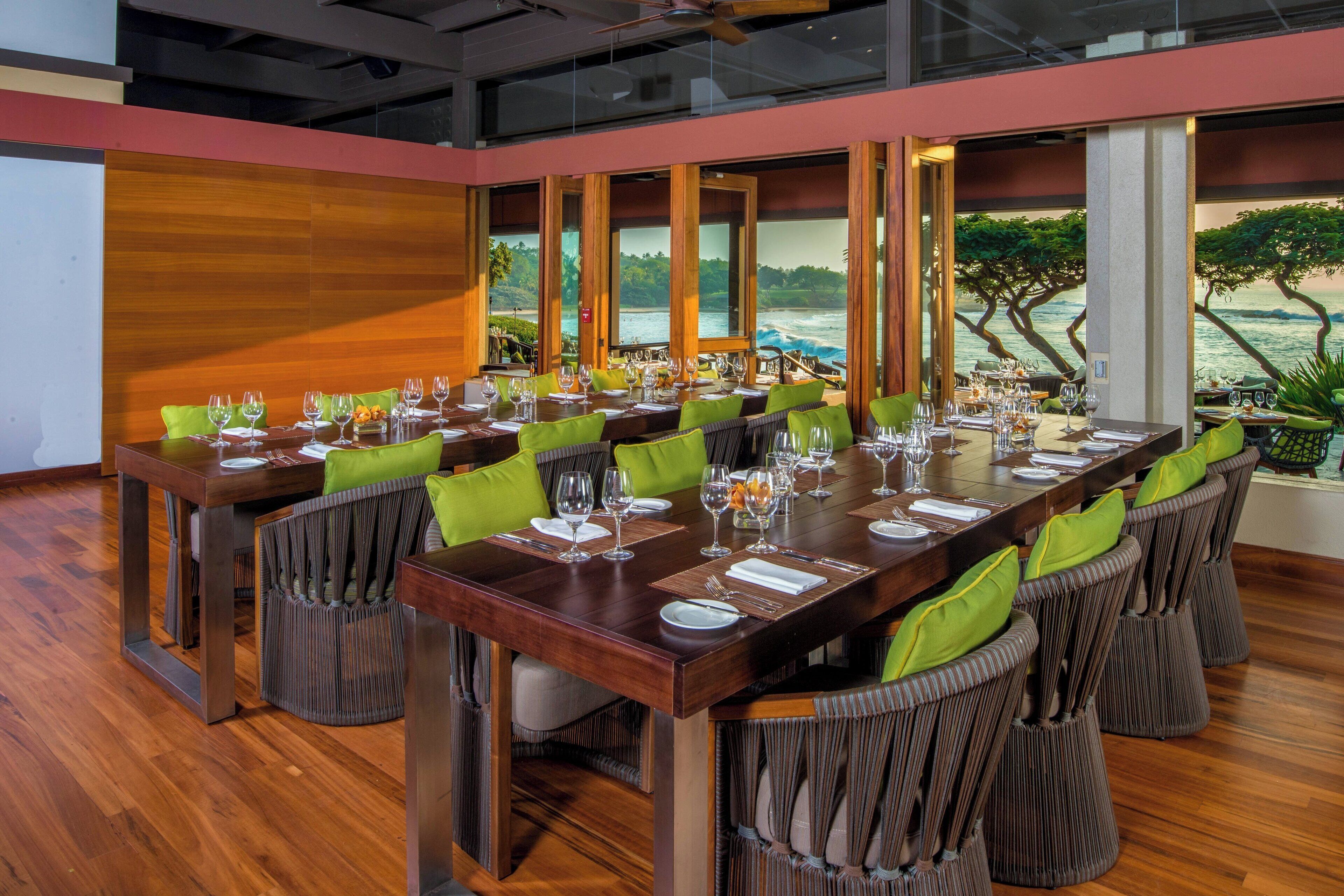 A dining area with a long wooden table set for a meal, surrounded by chairs with green cushions, large windows with a view of a body of water and greenery.