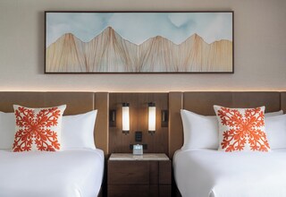 A hotel room with two beds, a nightstand, a clock, and a wall-mounted artwork depicting mountains.