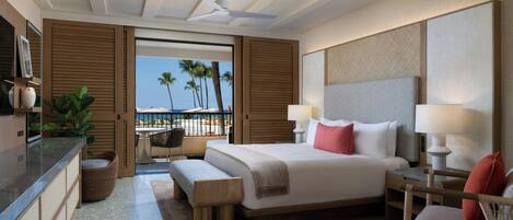 Quarto de hotel moderno com uma cama grande, uma escrivaninha, uma cadeira e vista para palmeiras e o oceano.