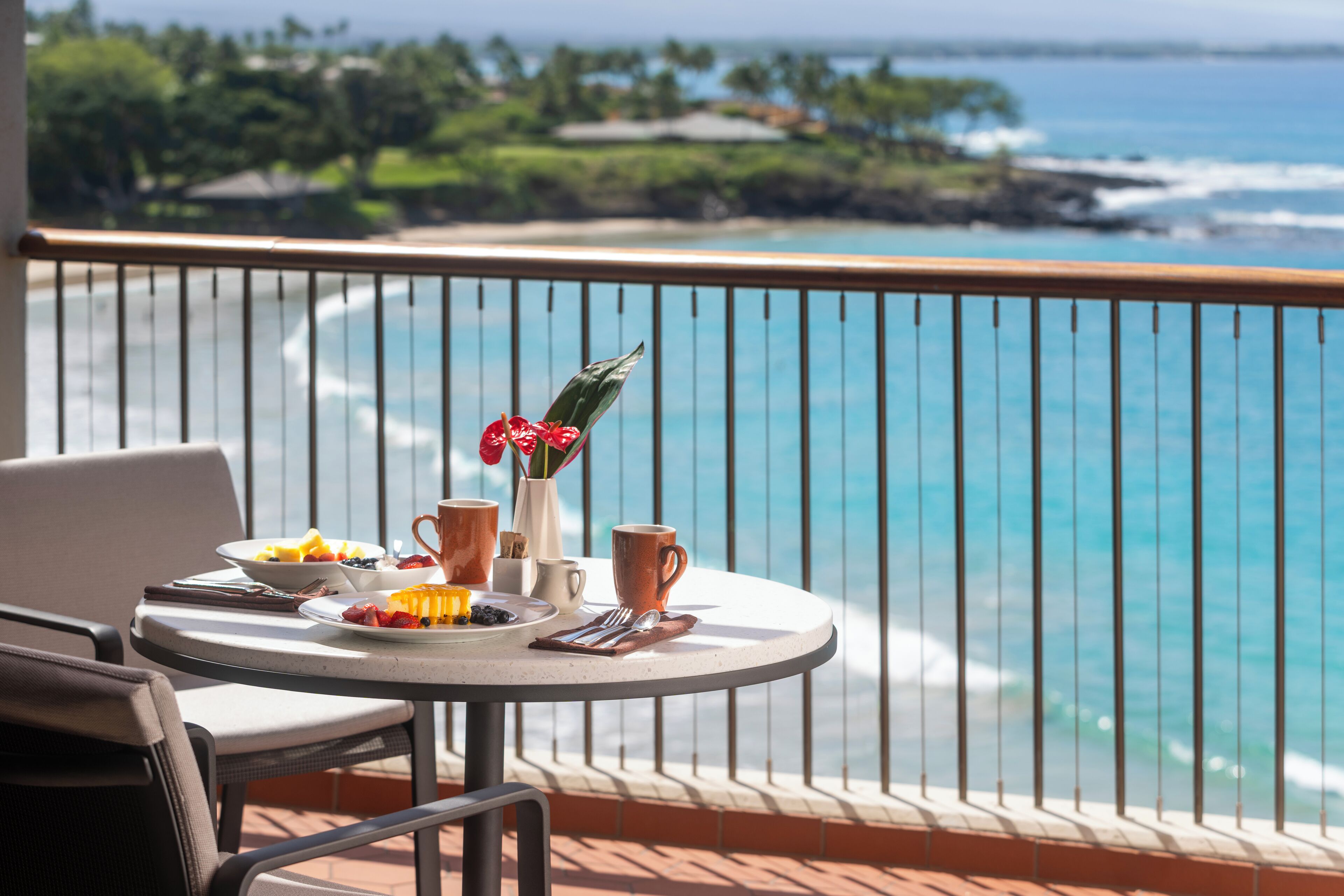 Une table ronde, dressée pour le petit-déjeuner, avec un vase de fleurs, donnant sur une plage où l’on aperçoit des vagues et une végétation luxuriante.