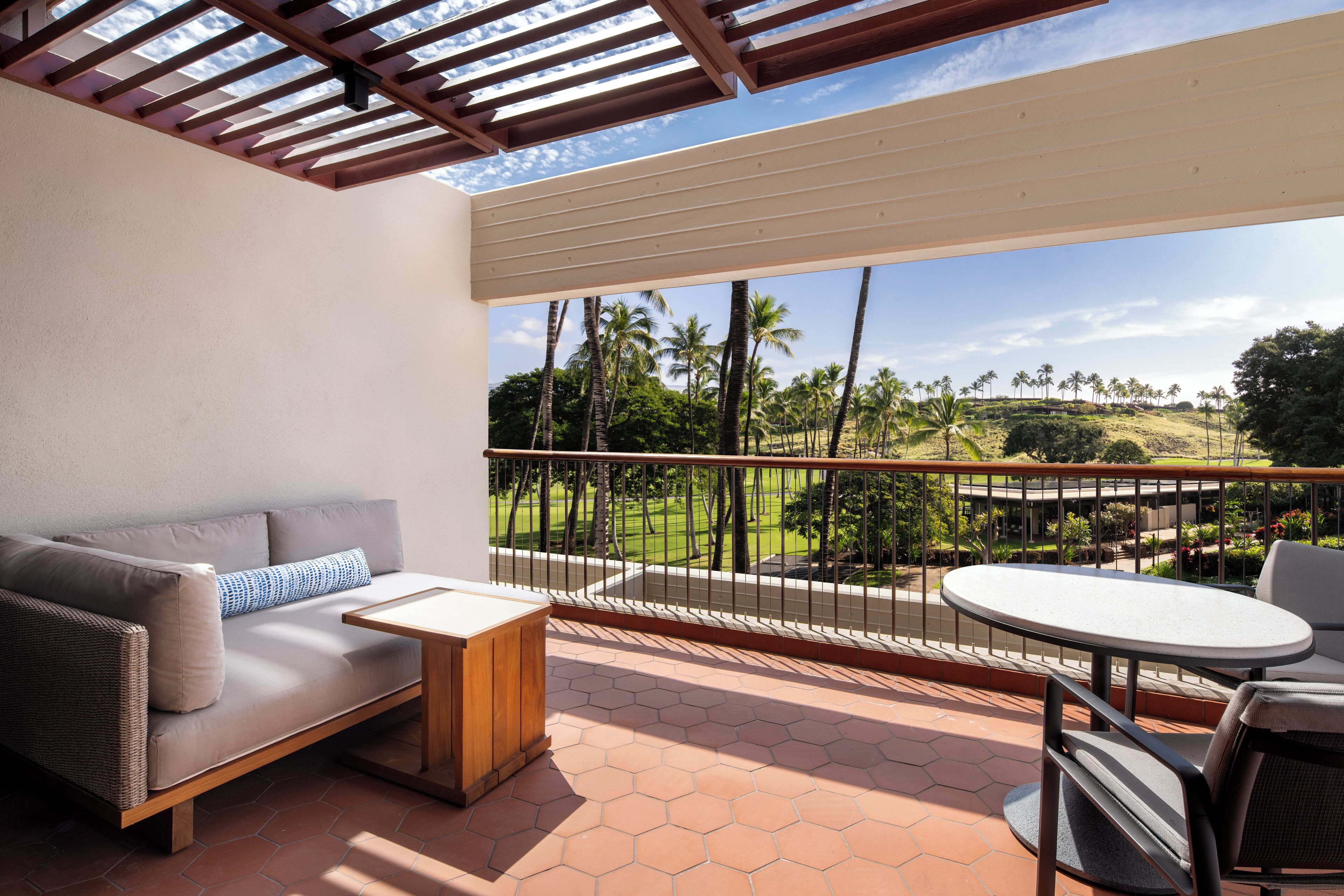 A balcony with a sofa, a small table, and a round table with chairs. The balcony overlooks a landscaped area with palm trees and a view of a hilly landscape.