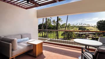 A balcony with a sofa, a small table, and a round table with chairs. The balcony overlooks a landscaped area with palm trees and a view of a hilly landscape.