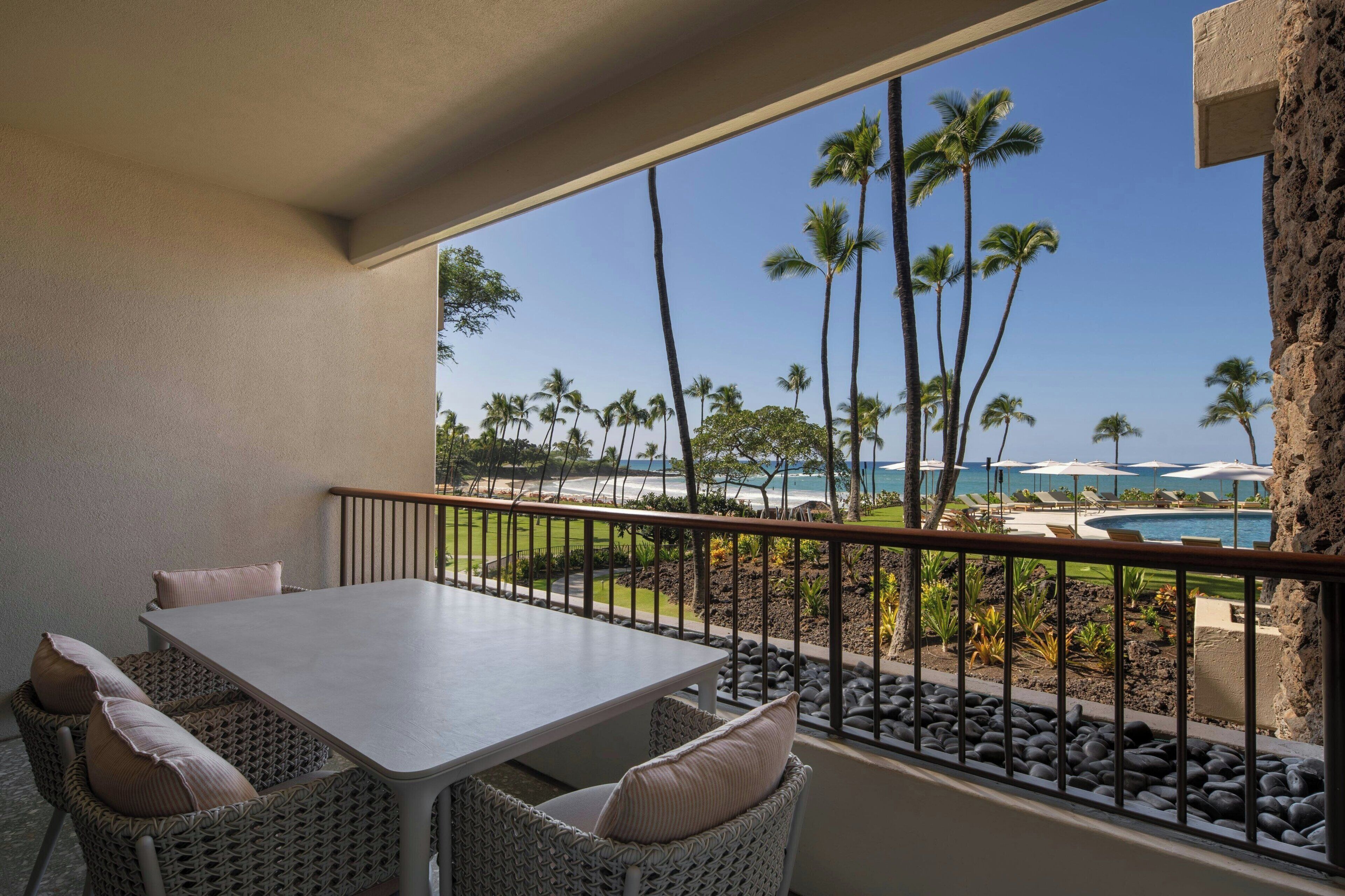 Un balcon avec une table et des chaises, donnant sur une piscine entourée de palmiers et offrant une vue sur l’océan.