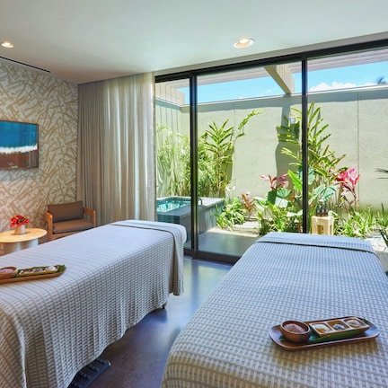 A hotel room with two beds, a small table with a vase, a chair, and a view of a garden through large windows.