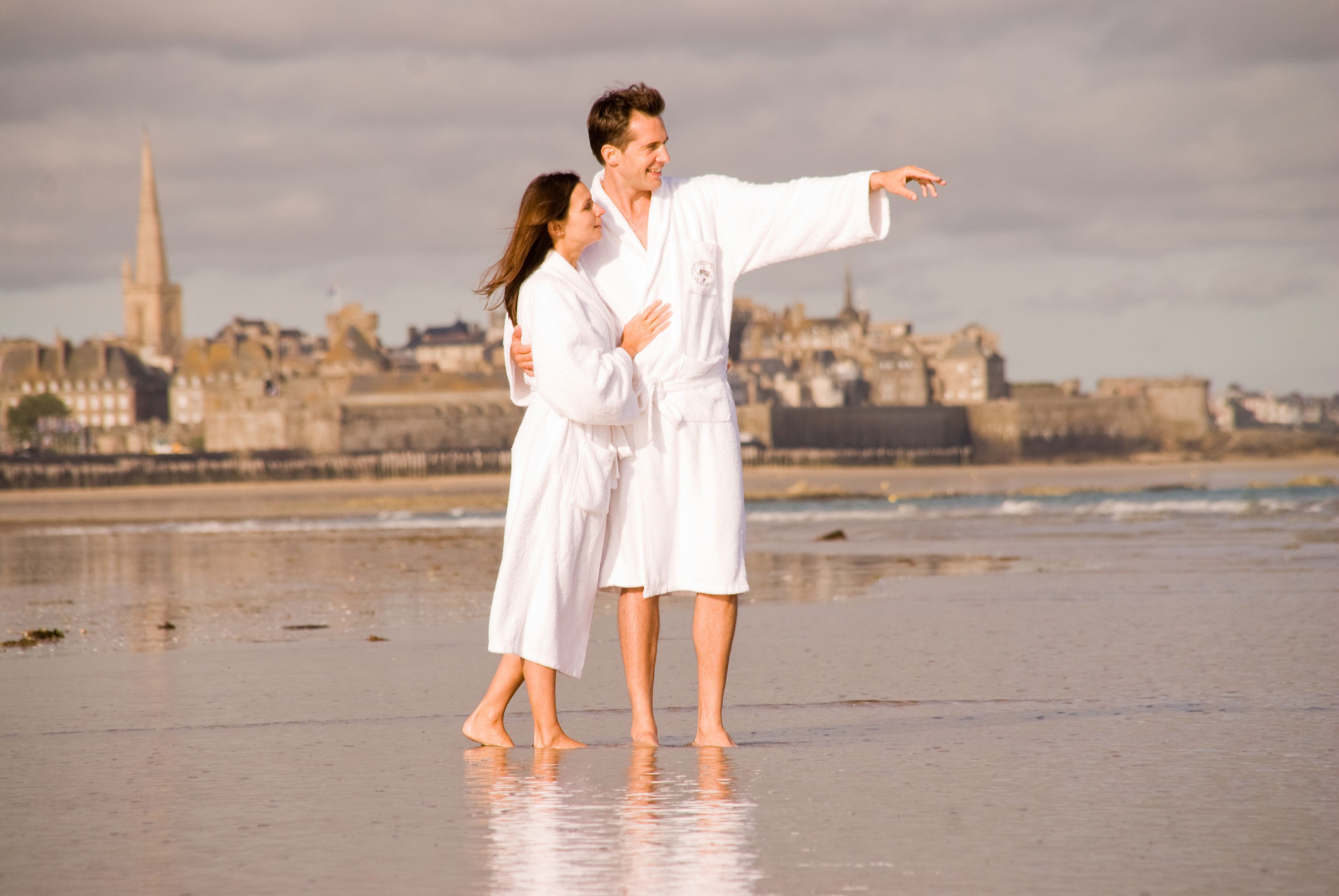 Una pareja vestida de blanco en una playa, con un paisaje urbano al fondo.