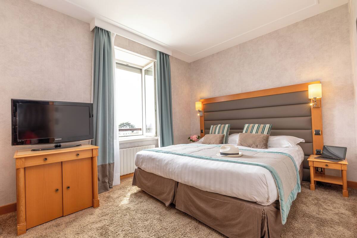 Een hotelkamer met een bed, een televisie op een houten kast, een laptop op een nachtkastje en een raam met gordijnen.