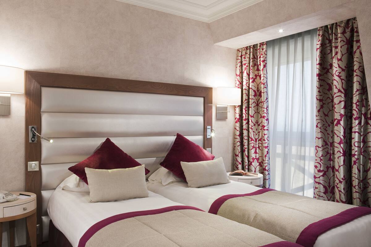 Hotelkamer met twee bedden, een houten hoofdbord, een nachtkastje met lamp en een raam met gordijnen met een patroon.