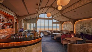 A bar area with a curved counter, shelves with bottles, and glasses. There are seating areas with tables and chairs, and a large window offering a view of the sea. - Grand Hôtel des Thermes (Saint-Malo)