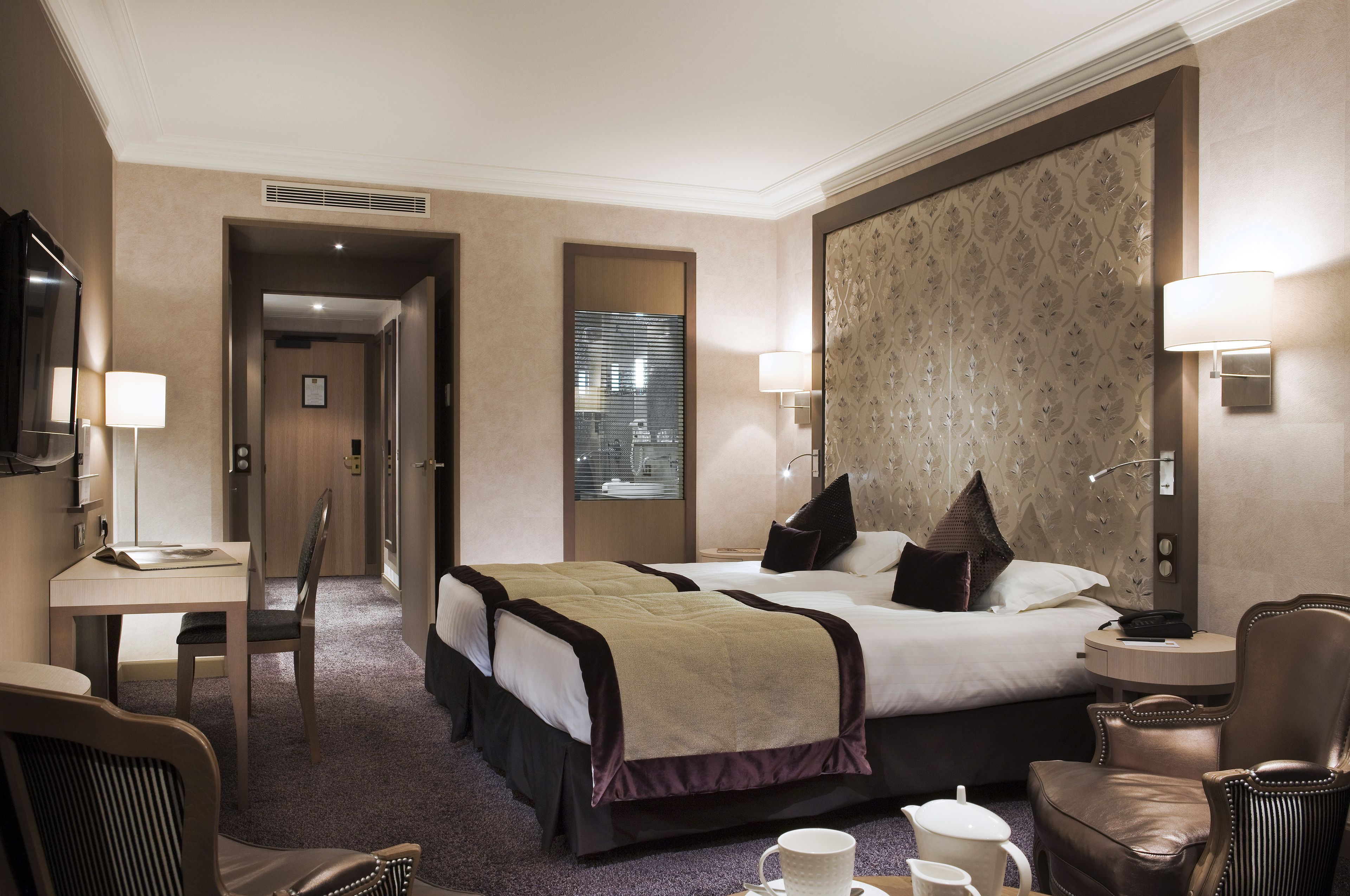 A hotel room with a large bed, a desk, a chair, and a small table with a cup and a pitcher.