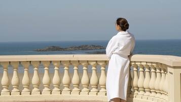 Une personne vêtue d’une longue robe blanche se tient sur un balcon orné d’une balustrade, d’où elle domine un plan d’eau et des formations rocheuses.