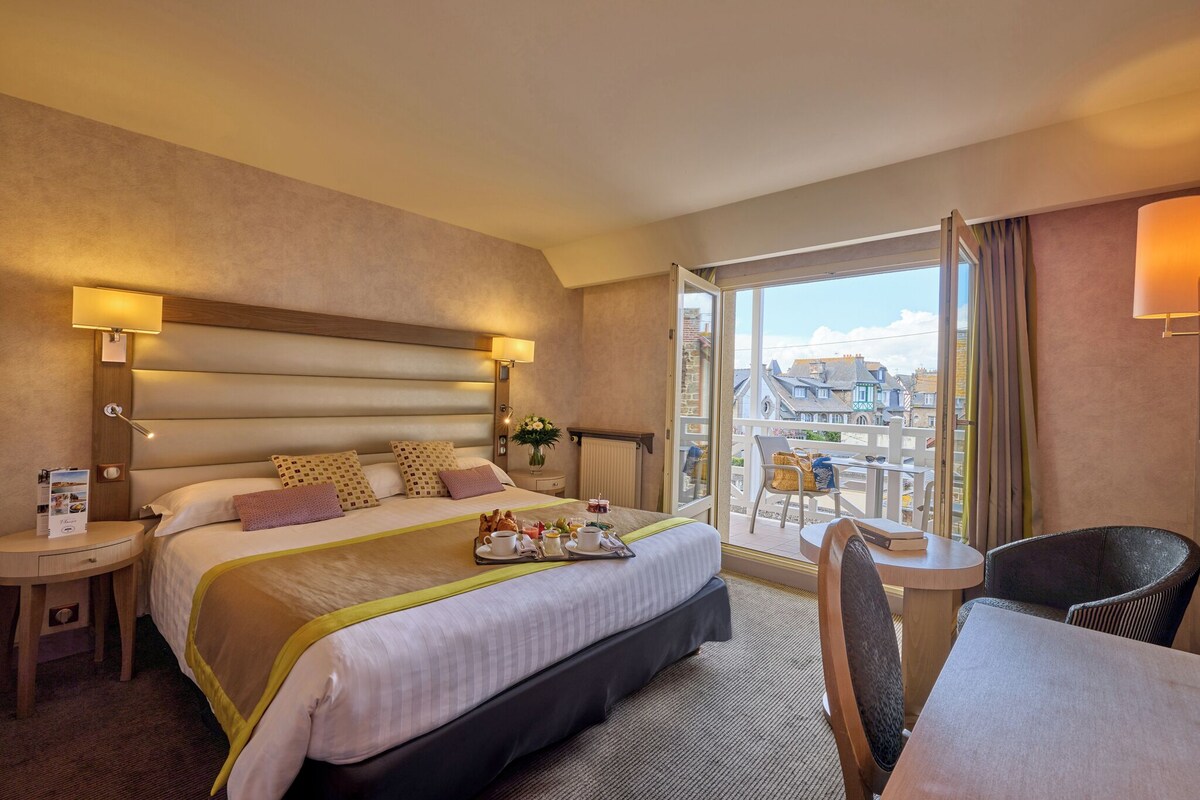 A hotel room with a large bed, bedside tables, a dining area, and a balcony with a city view.
