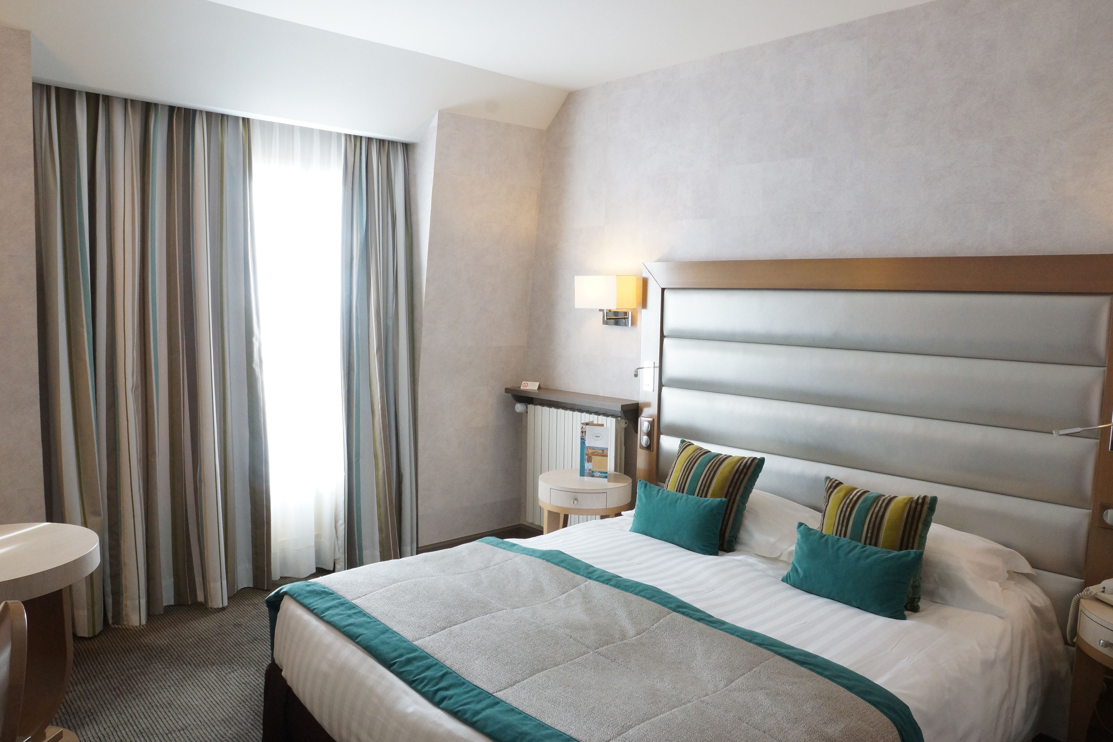 A hotel room with a large bed, a bedside table, a lamp, and a view of the outside through curtains.