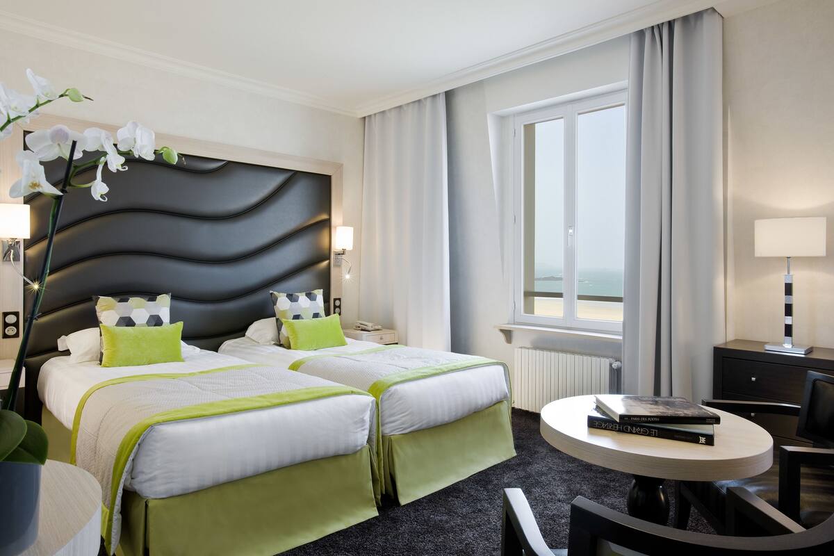 Een hotelkamer met twee bedden, een ronde tafel, een stoel, een bureau en een raam met gordijnen.