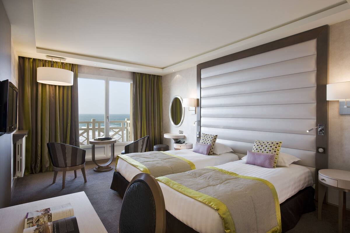 Een hotelkamer met twee bedden, een bureau, een stoel, een tafeltje, een televisie en uitzicht op zee.