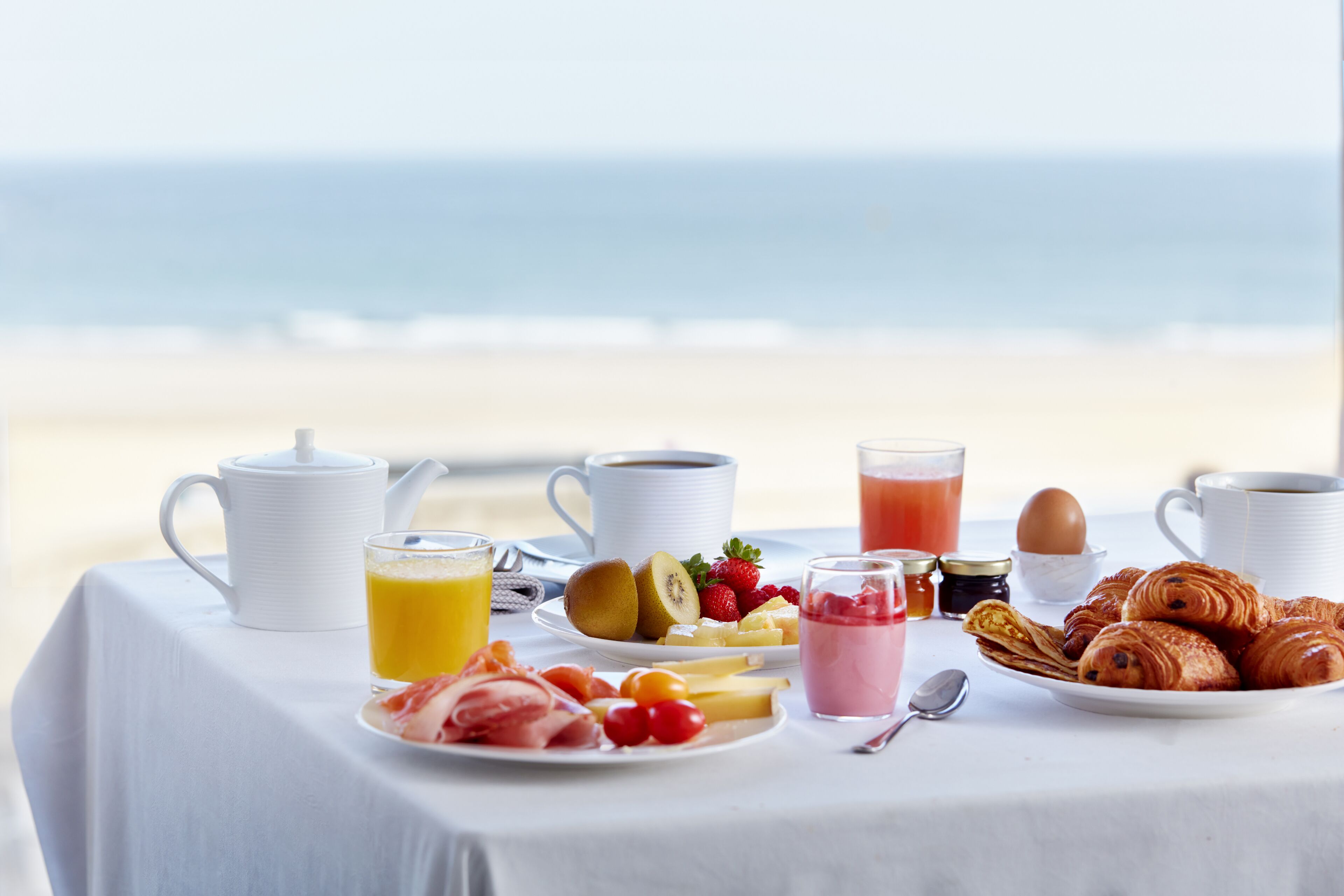 A breakfast table with various food items and drinks on a beach setting.