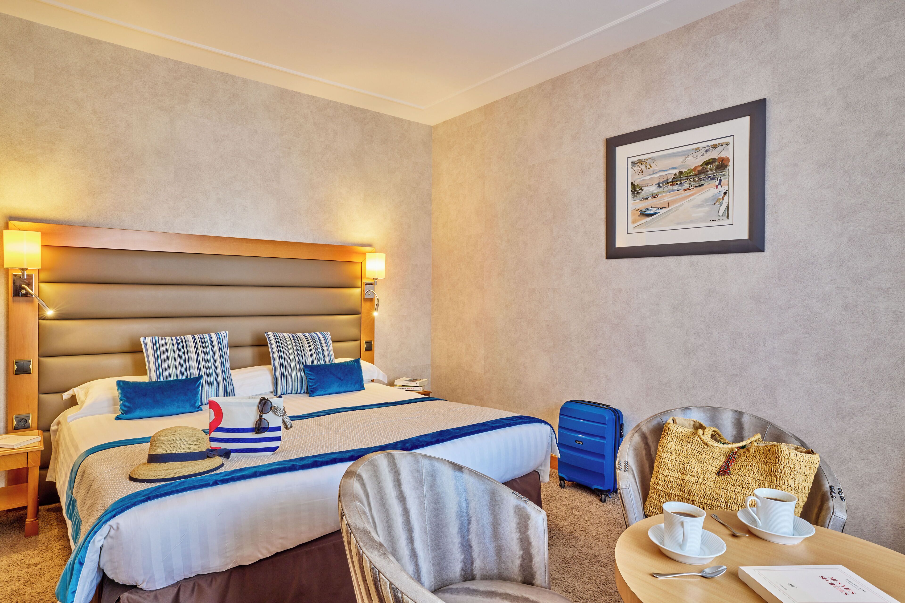 A hotel room with a large bed, a bedside table, a chair, a table with a cup and saucer, and a suitcase.