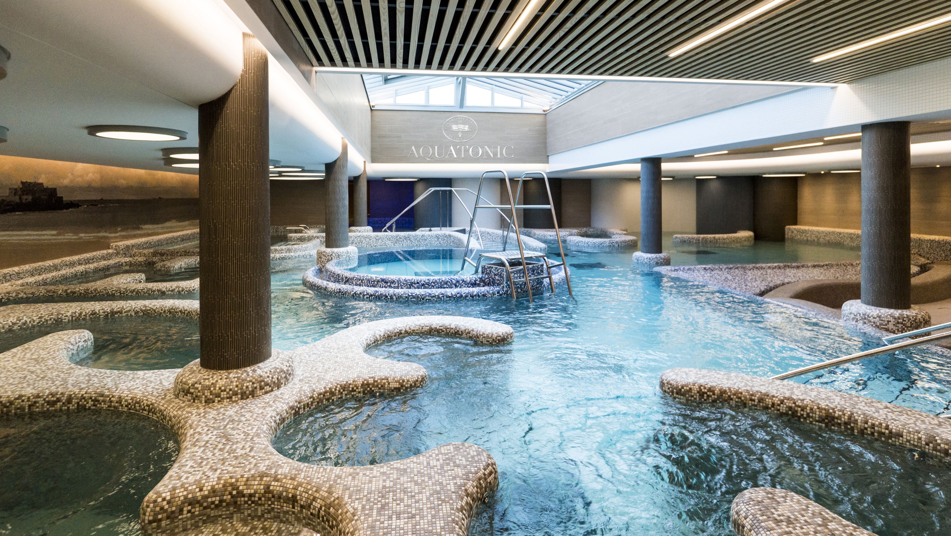 Une piscine intérieure aux murs recouverts de mosaïques, avec un escalier central et un panneau indiquant « AQUATONIC ».
