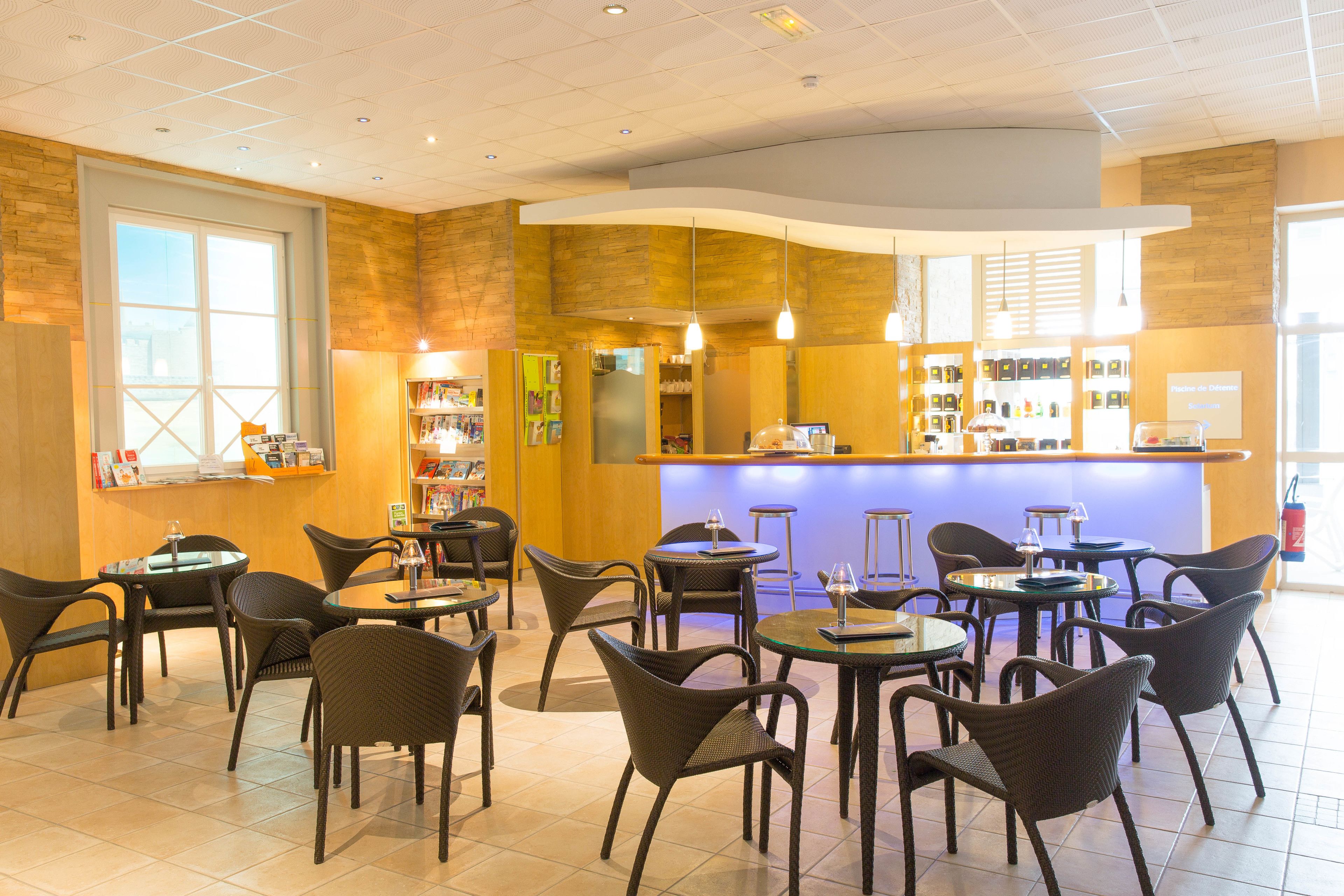 A cafe with multiple tables and chairs, a counter with various items, and a large window on the left.