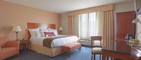 Une chambre d’hôtel avec un grand lit, un bureau avec une chaise, une petite table avec une lampe et une fenêtre avec des rideaux.