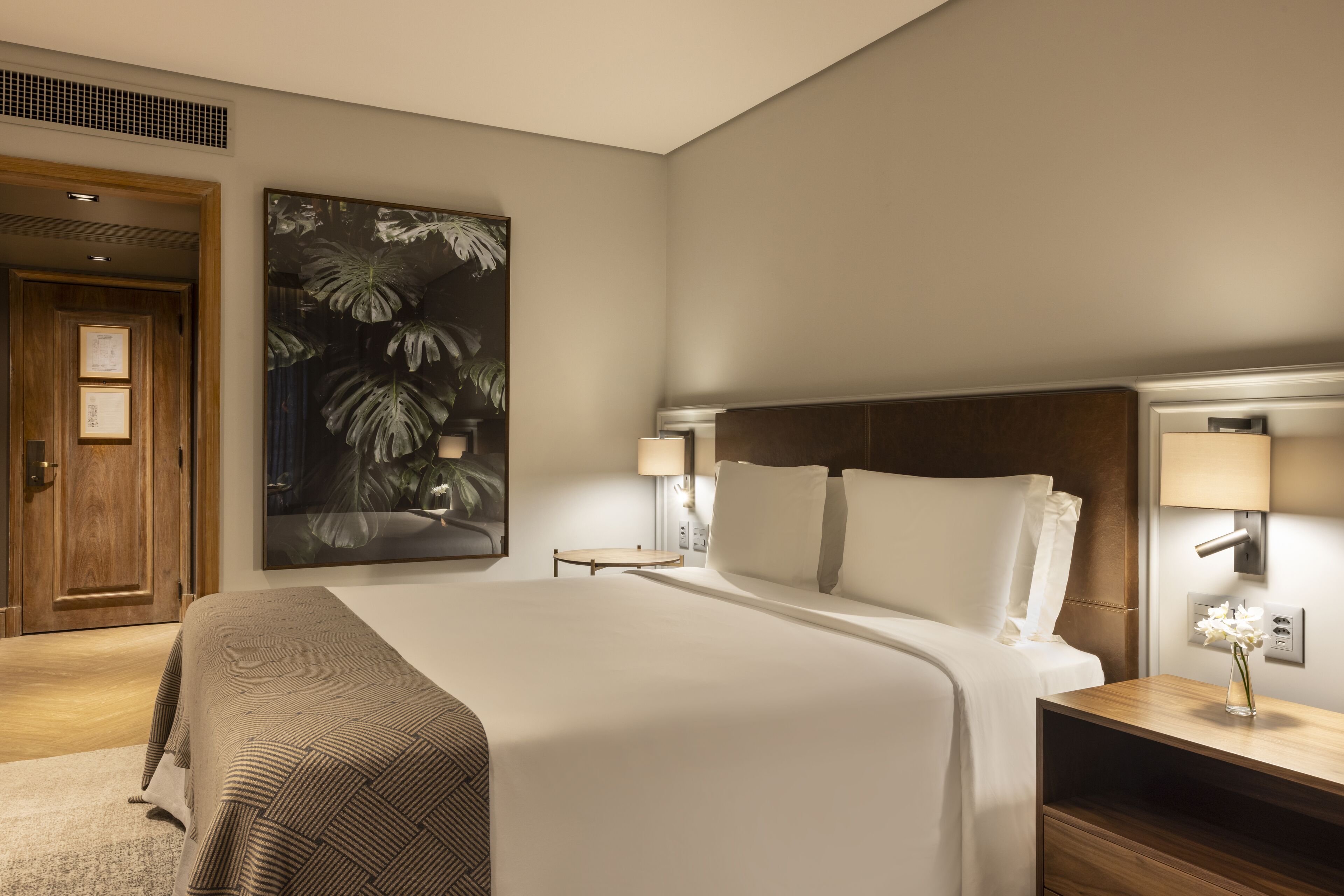 Habitación de hotel con una cama grande, una mesita de noche con lámpara, una lámpara de pared y una obra de arte con motivos de plantas tropicales.