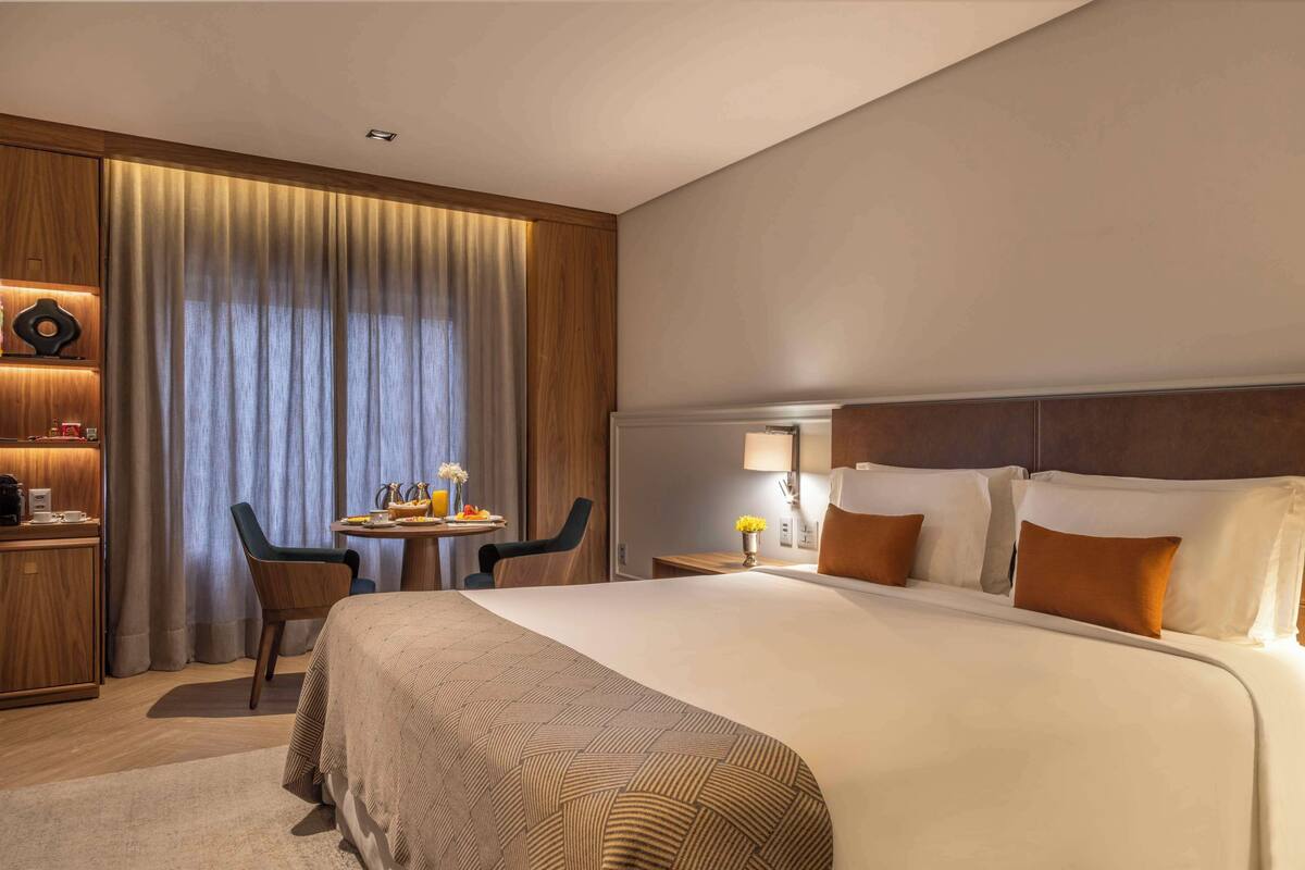 Een hotelkamer met een groot bed, een bureau met stoel, een tafeltje met lamp en een raam met gordijnen.