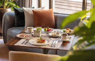 A dining table set with plates, cutlery, and a variety of food items, positioned in a cozy room with a sofa and cushions.