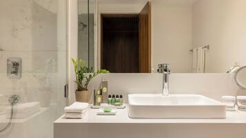 A modern bathroom with a large white sink, a glass shower enclosure, a wooden door, and a mirror reflecting the room.