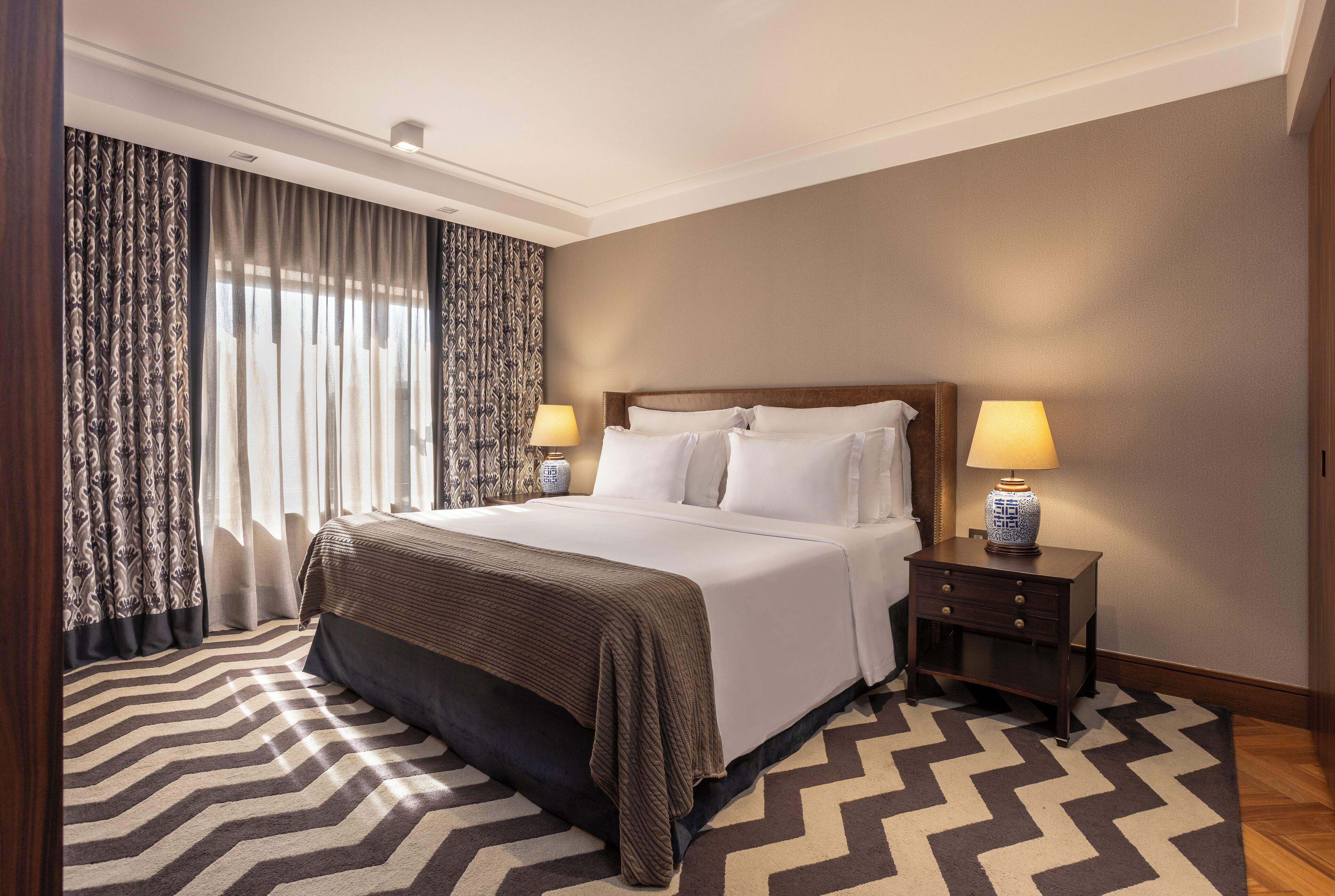 Camera d'albergo con un letto grande, due comodini con lampade, un tappeto decorato e vista sull'esterno attraverso tende leggere.