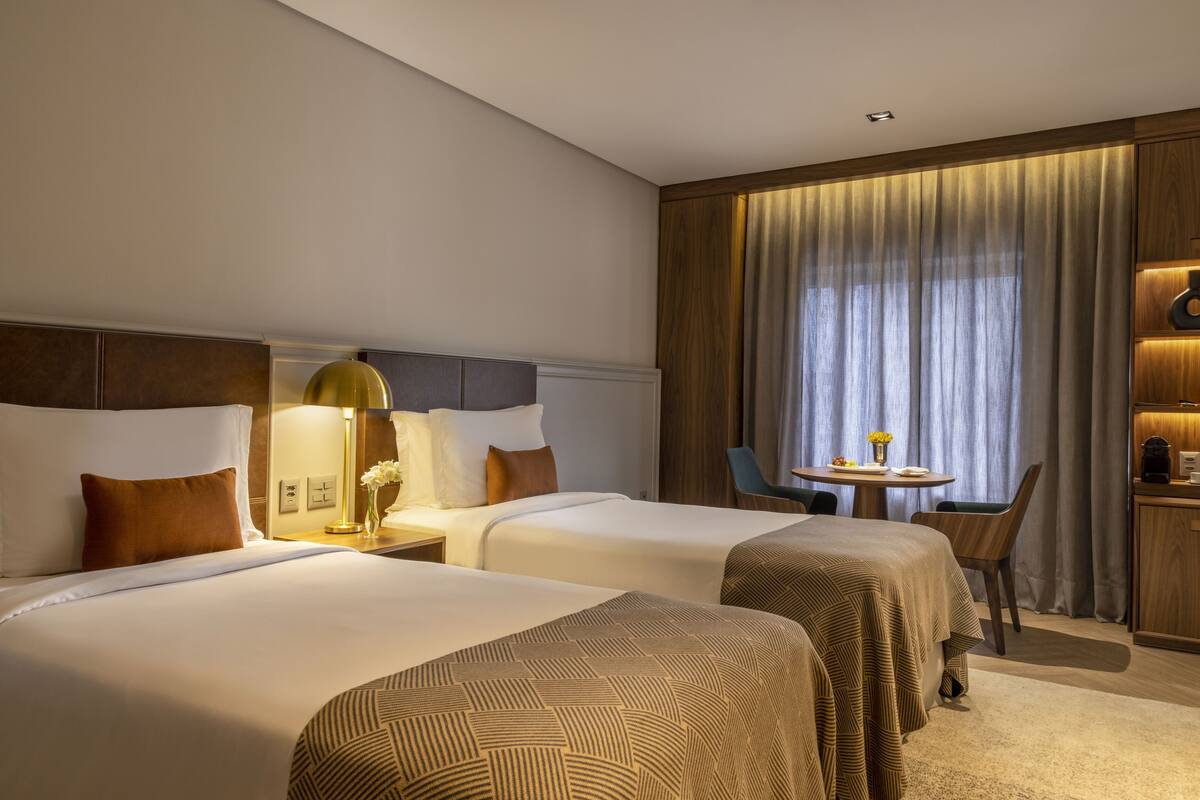Een hotelkamer met twee bedden, een kleine tafel met stoel, een bureau met lamp en een groot raam met gordijnen.
