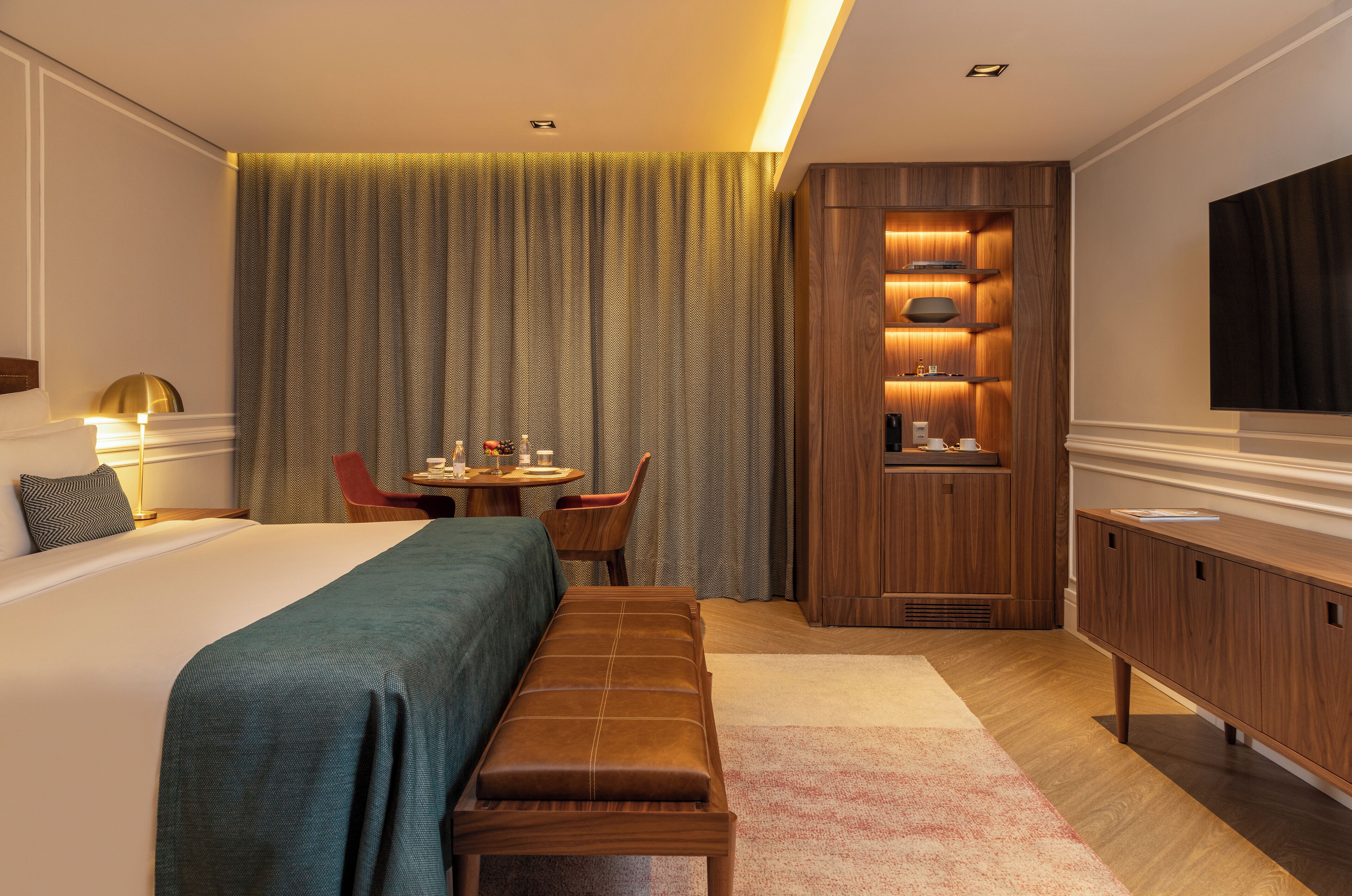 A hotel room with a large bed, a bench, a desk with a chair, a TV, and a wooden cabinet.