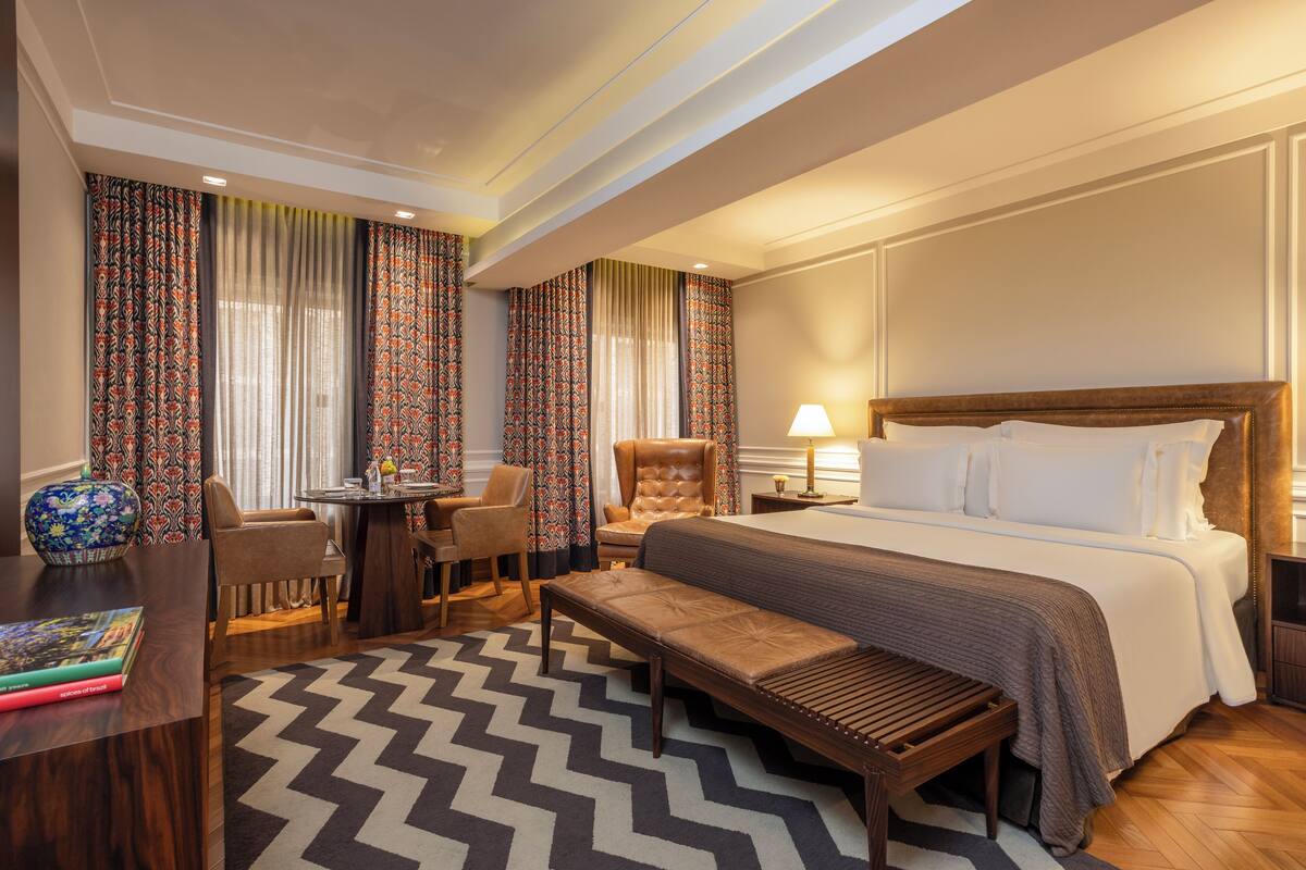 Een hotelkamer met een groot bed, een bureau met stoel, een bankje en een kleine tafel met vaas.
